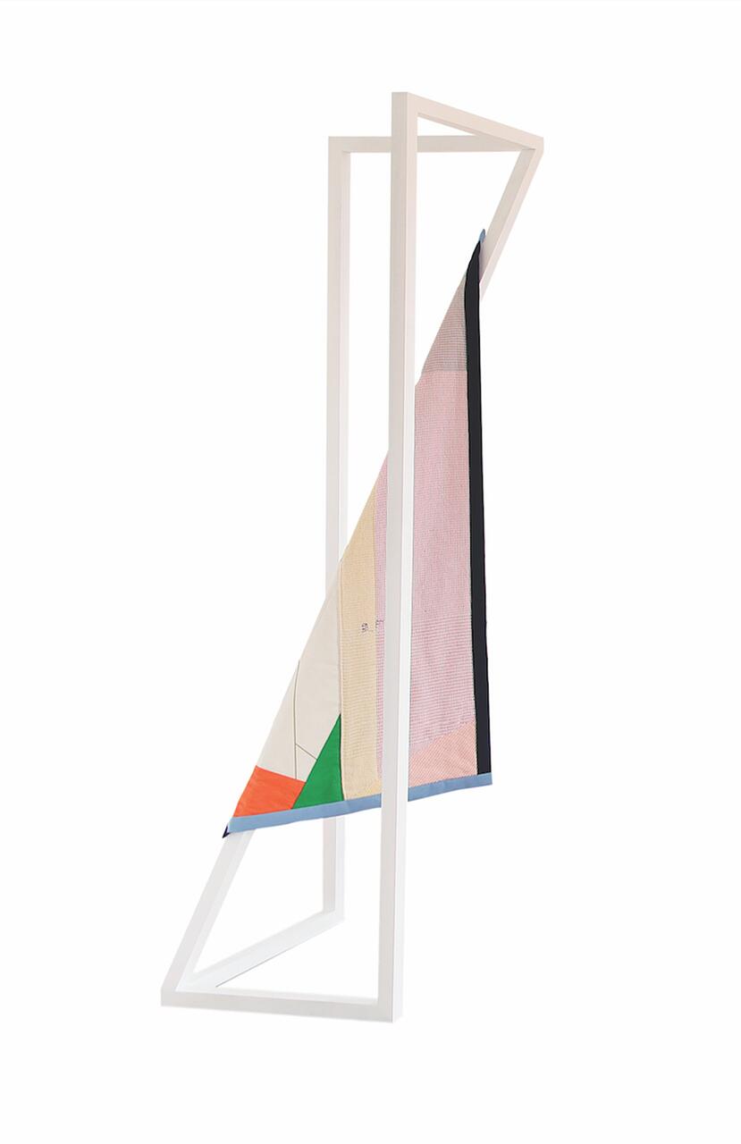 Geometric sculpture with a white frame, holding a colorful fabric piece. The fabric features red, green, blue, and beige sections, suspended diagonally within the structure.