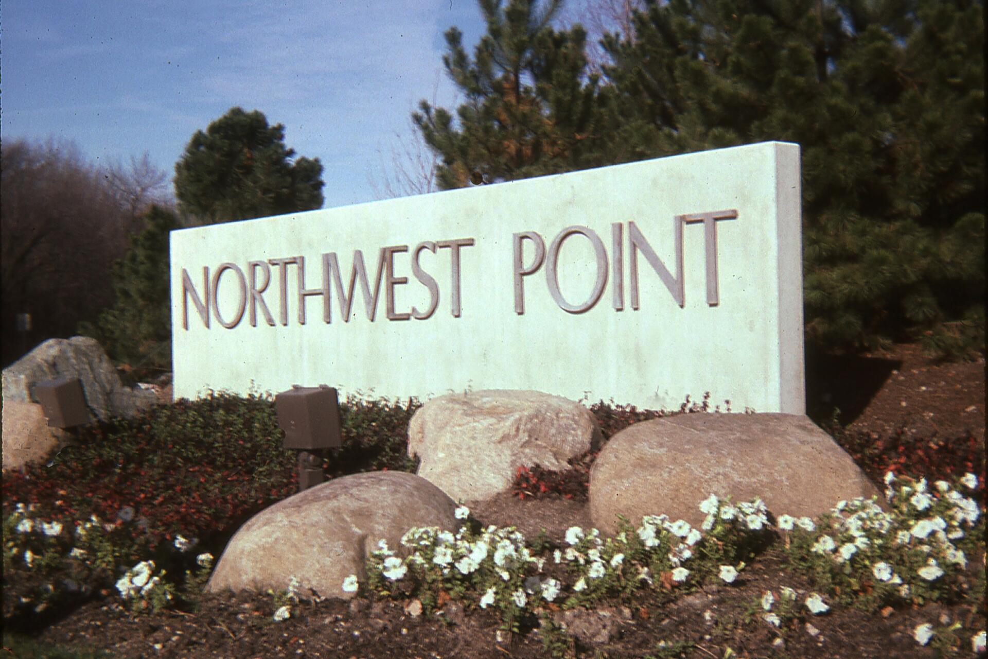 Stone sign reading "Northwest Point" surrounded by rocks, shrubs, and white flowers, with evergreen trees in the background.