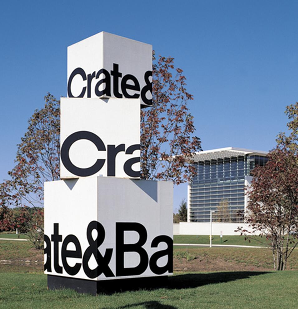 Three stacked white cubes with bold black lettering, "Crate & Barrel," against a blue sky. Nearby, a modern building with a glass facade and trees with autumn leaves create a serene setting.
