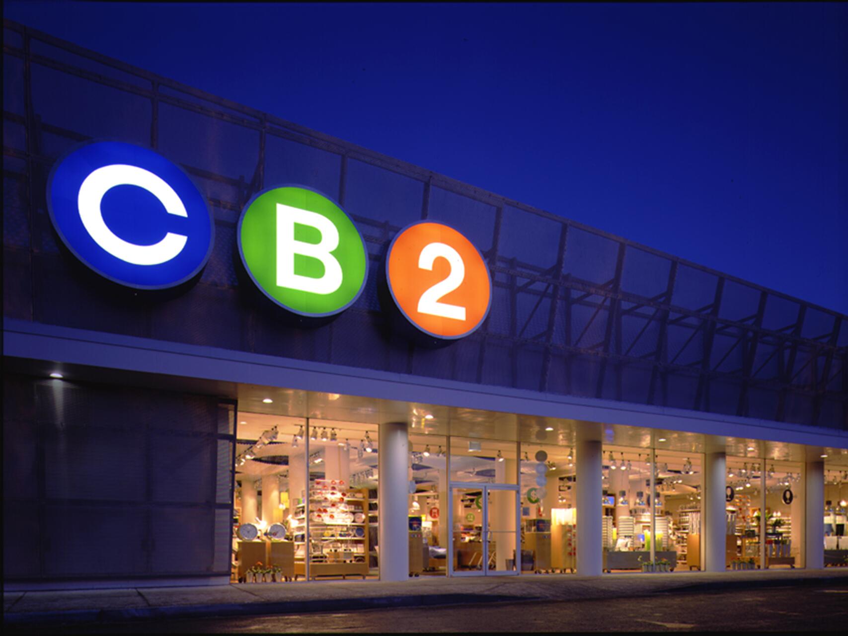 Storefront with illuminated "CB2" logo in blue, green, and orange circles against a twilight sky. Brightly lit interior displays furniture and home decor through large glass windows.