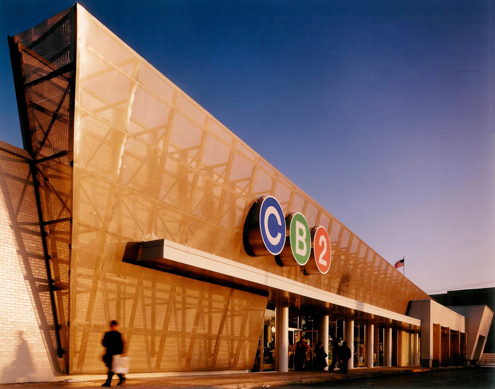 Modern store exterior with a large glass facade, featuring the letters "C", "B", and "2" in colorful circles. A person is walking by under a clear blue sky.