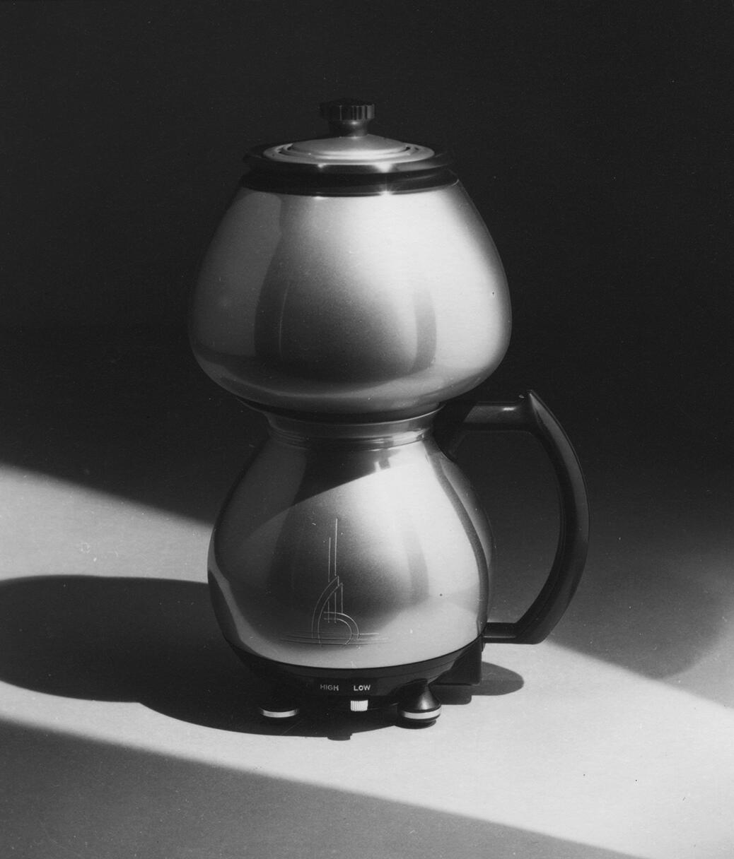 Stainless steel coffee maker with a curvy design, black handle, and lid, set against a dark backdrop with dramatic lighting.