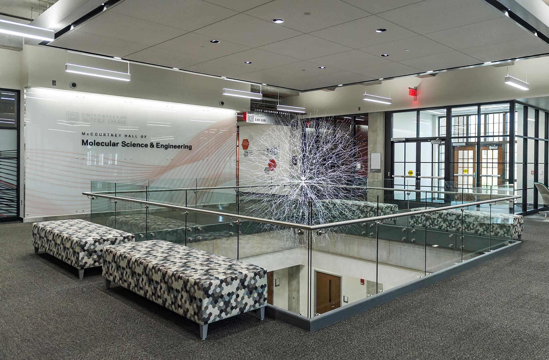 Modern hallway in McCourtney Hall of Molecular Science & Engineering, with glass railings, geometric-patterned benches, and a large abstract sculpture featuring a network of white lines.