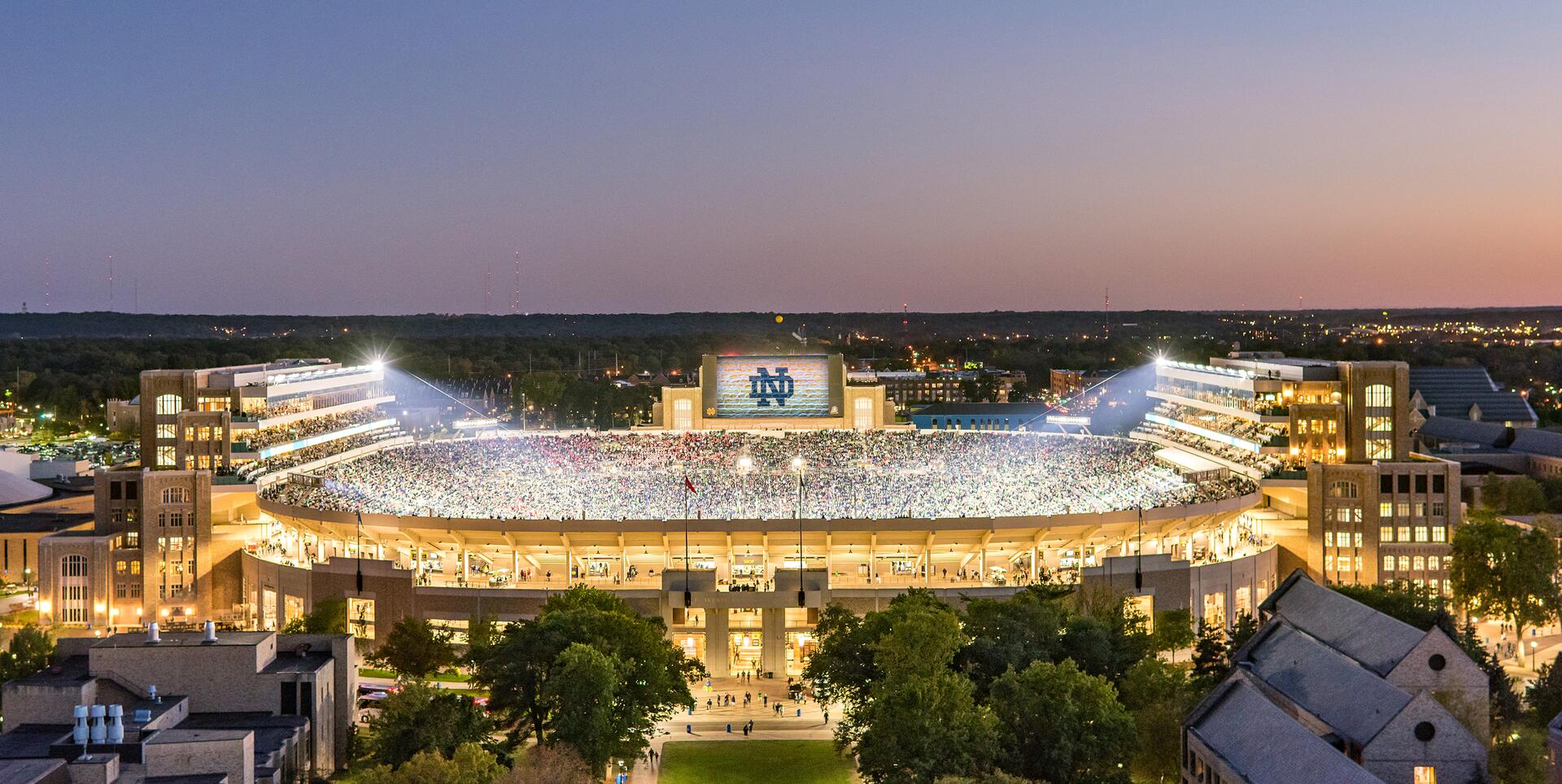 Illuminated football stadium at dusk, packed with fans. A large screen displays a logo, surrounded by lit stands and a twilight sky. Trees and buildings visible in the foreground.