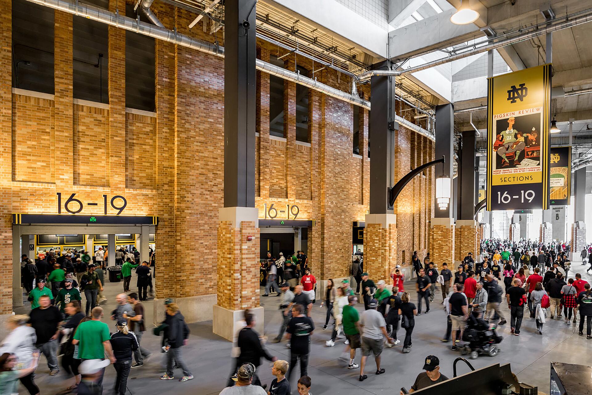 Crowd of people walking through a stadium concourse with brick walls and section signs labeled 16-19. A large banner hangs overhead displaying the same section numbers and a colorful illustration.