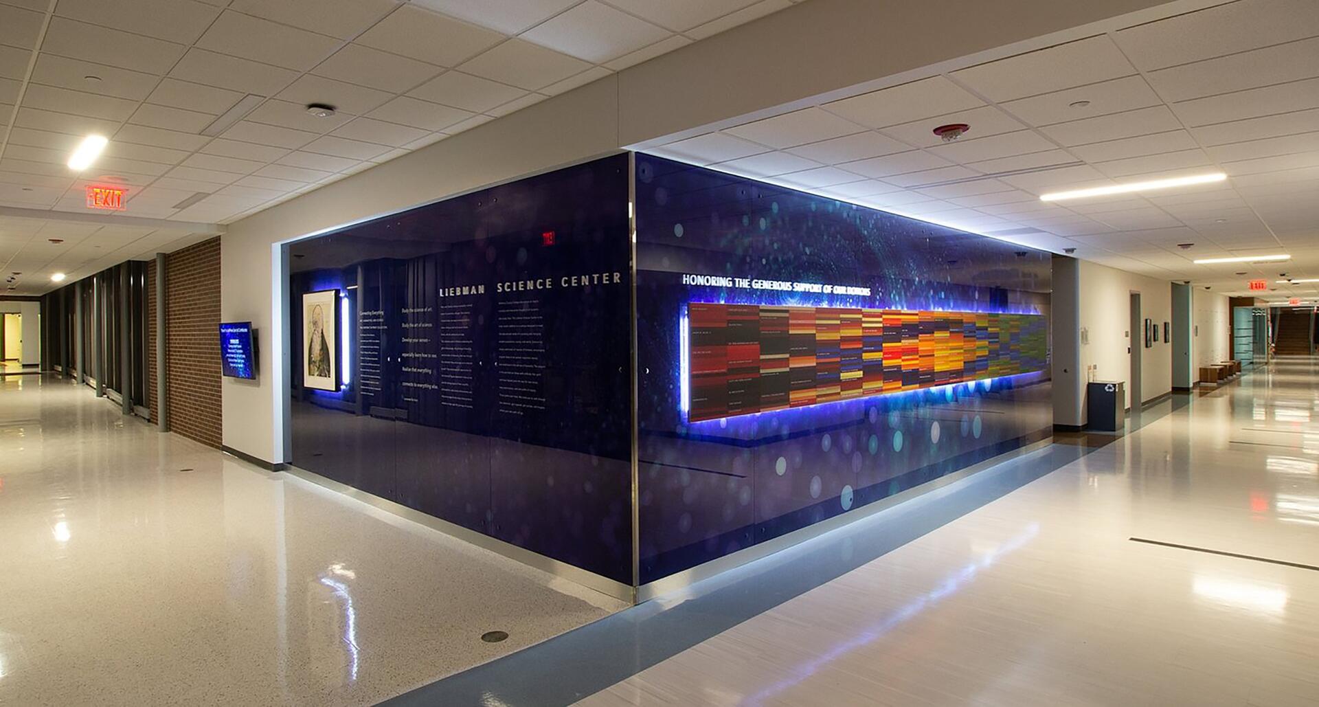 Hallway with polished floors, showcasing a corner plaque with colorful panels and text, titled "Liebman Science Center." Ceiling lights and an exit sign illuminate the space.