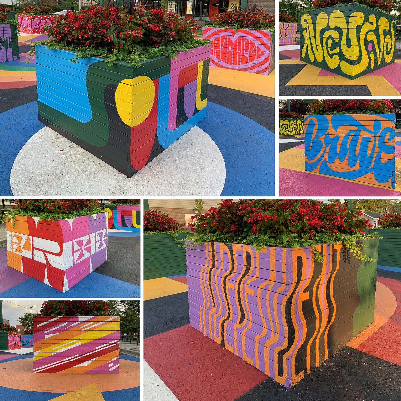 Colorful planters in a public square, each decorated with vibrant geometric designs and stylized letters. Red flowers and greenery spill over the tops. The ground features multicolored circular and geometric patterns.