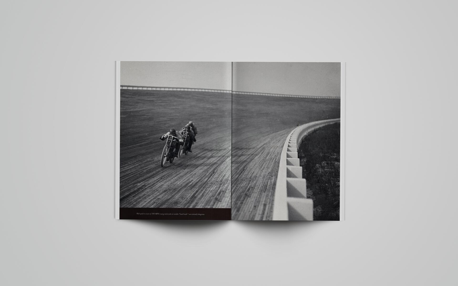 Black and white photo of two motorcyclists racing on a steep, curved wooden track, with a white barrier on the right. The riders are leaning into the curve, creating a sense of speed and intensity.