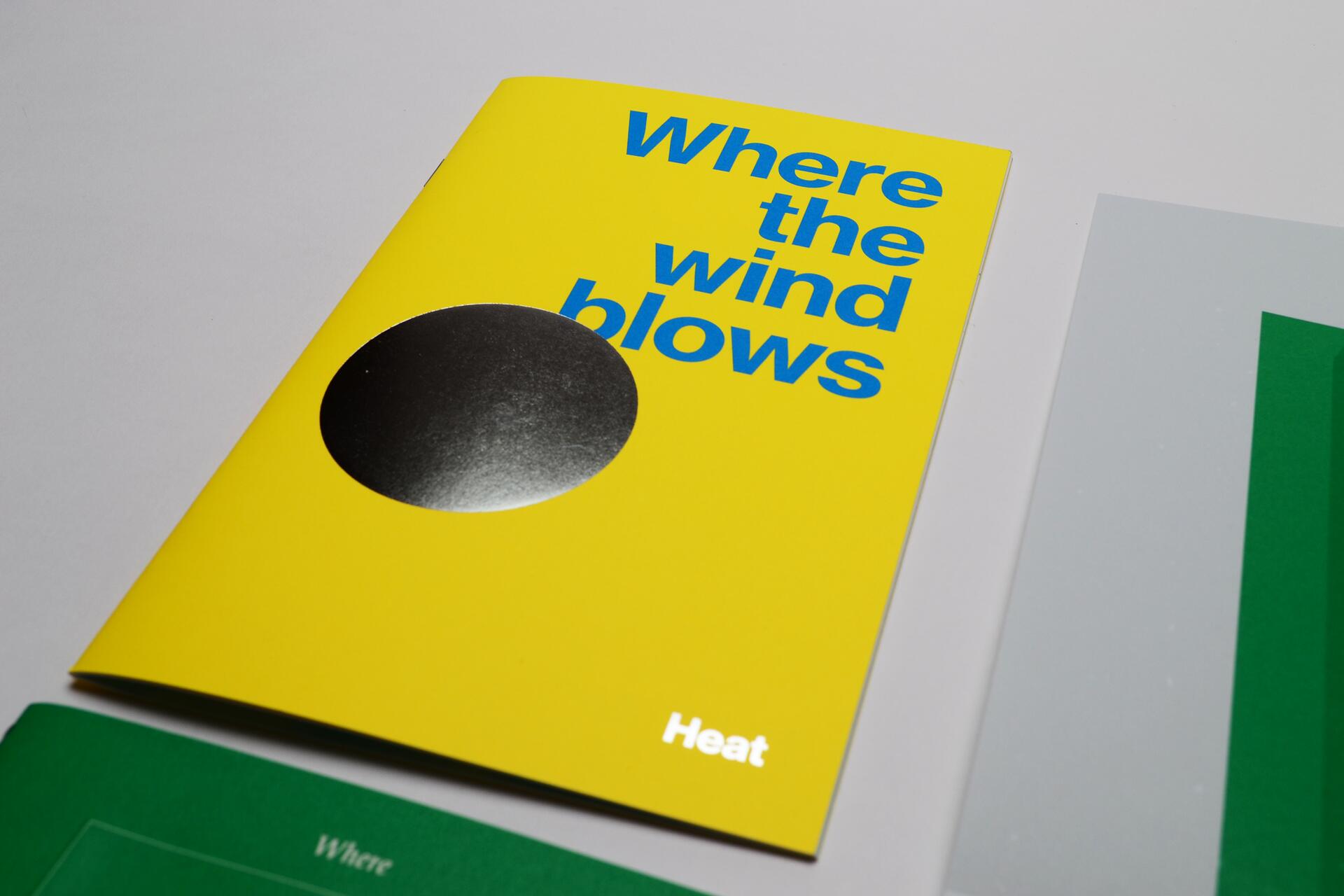 Bright yellow booklet with blue text, "Where the wind blows," and a black circle. The word "Heat" appears at the bottom right. Partially visible green and gray papers beneath.